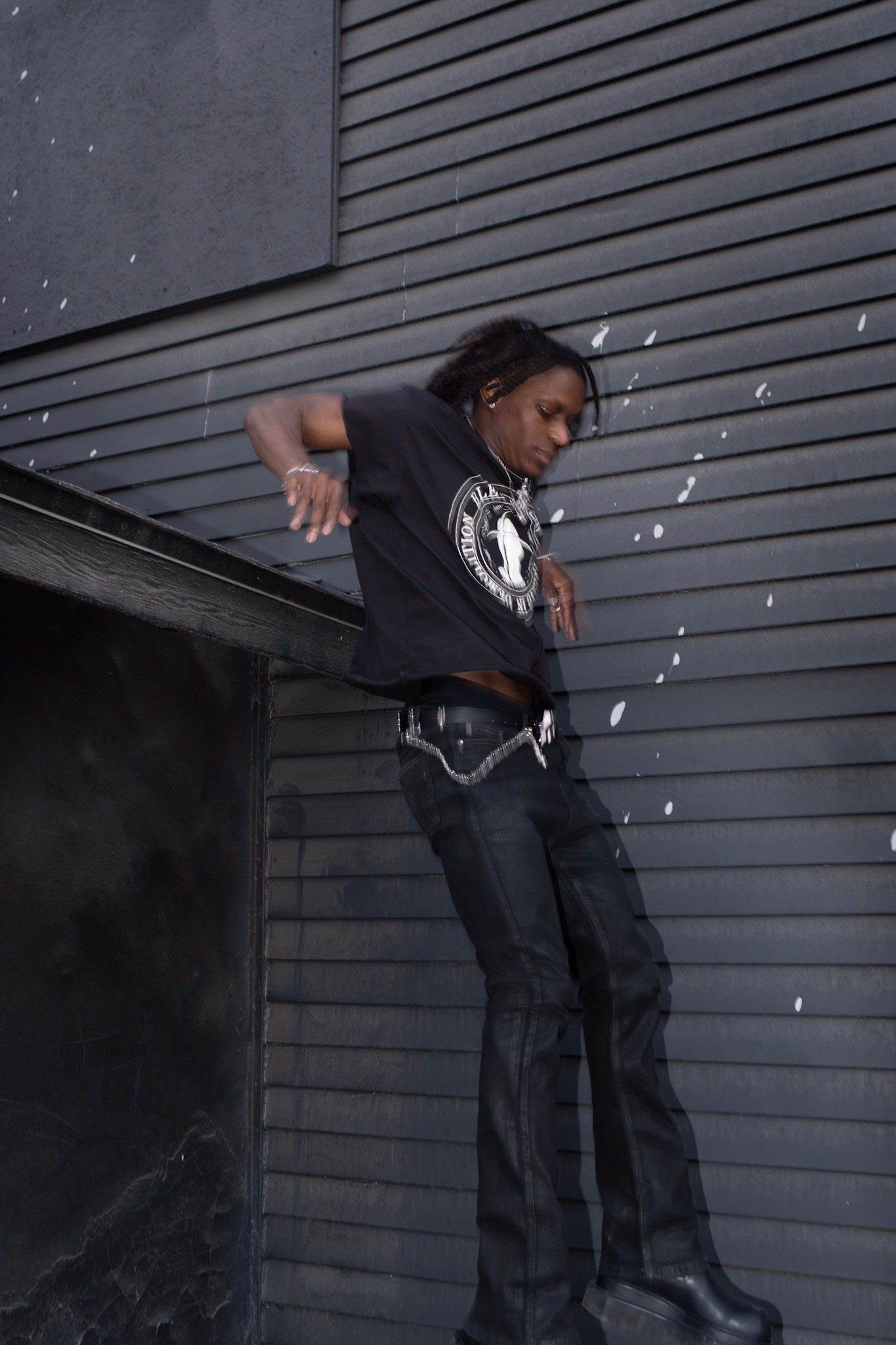 Person wearing a black t-shirt and jeans leaning against a dark gray wall.